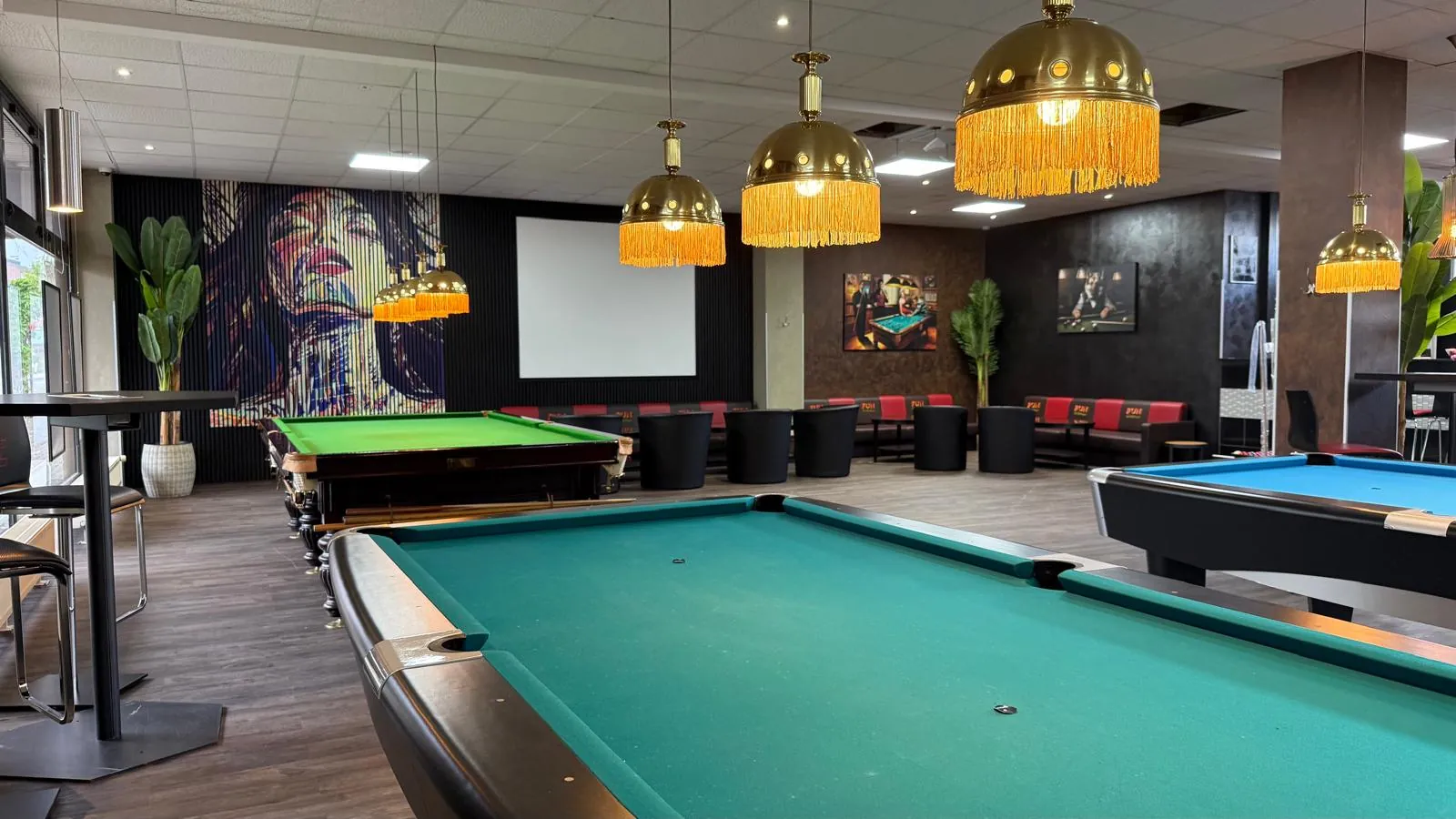 Saal von Billard Voerde mit Pooltischen unter goldenen Fransenlampen, links der Snookerbereich, hinten Leinwand und Sitzecken